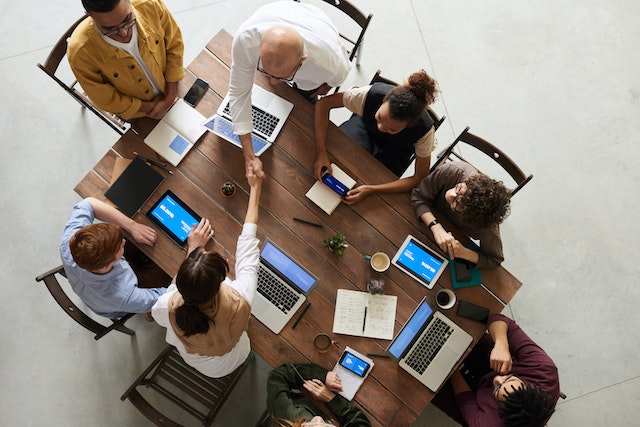 Eight people seated at a large wooden table having a meeting with their phones and laptops